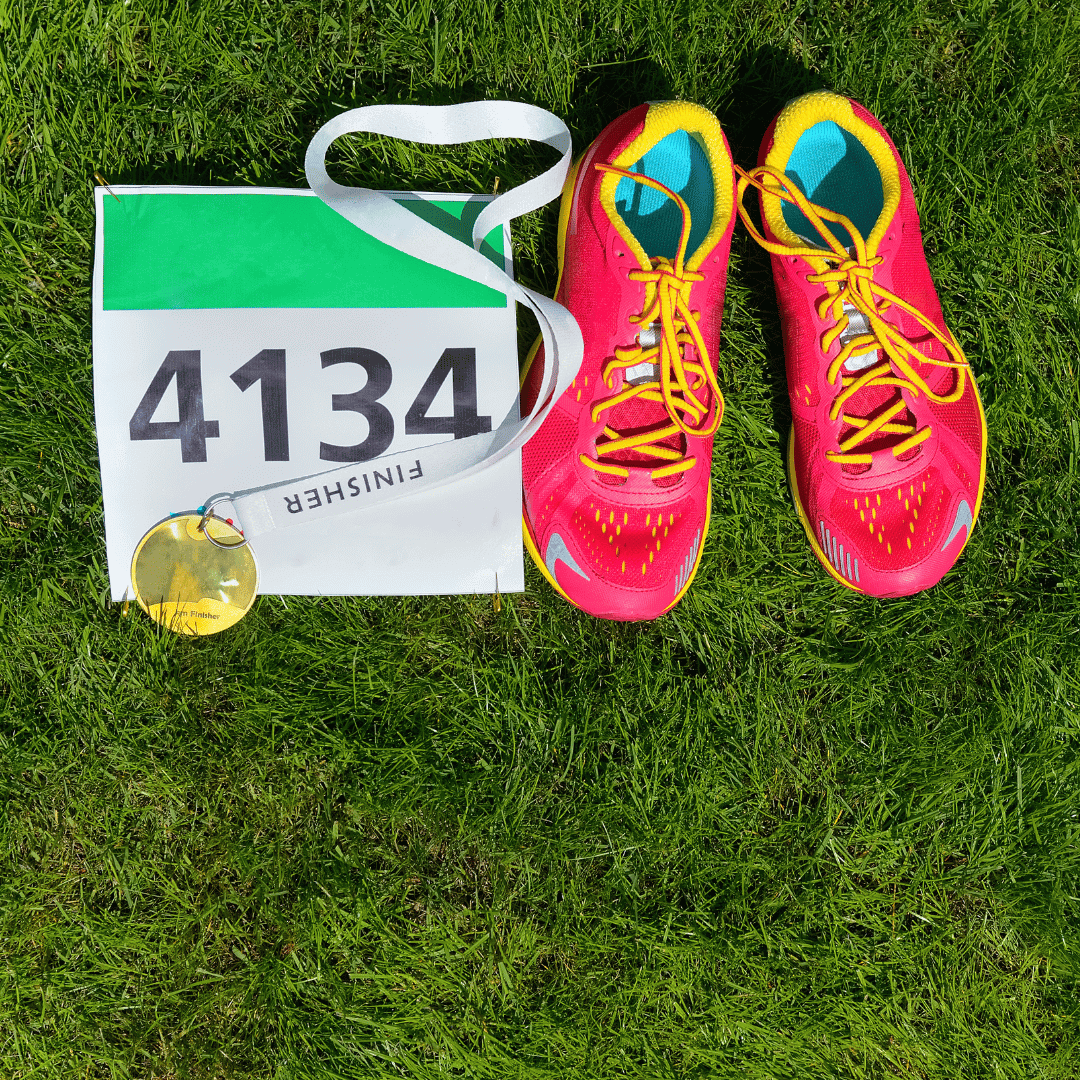 on a patch of grass there's a marathon racing bib to the left, gold medal on top and a red pair of sneakers to the right.